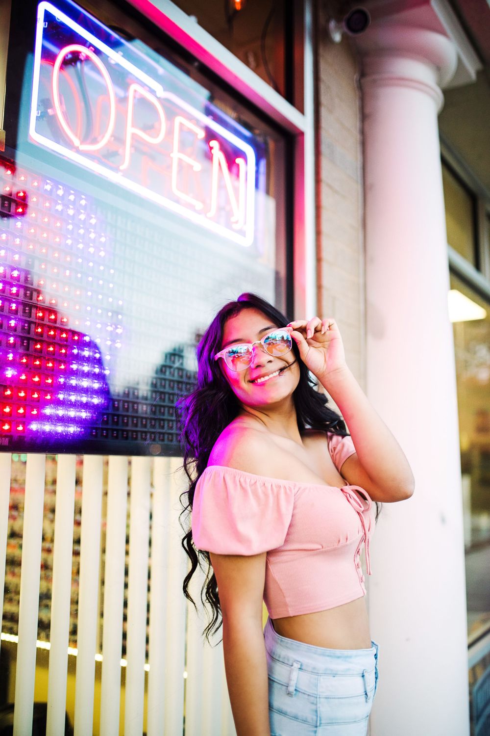 Smiling young woman in pink crop top adjusts sunglasses beside a colorful neon open sign at night.