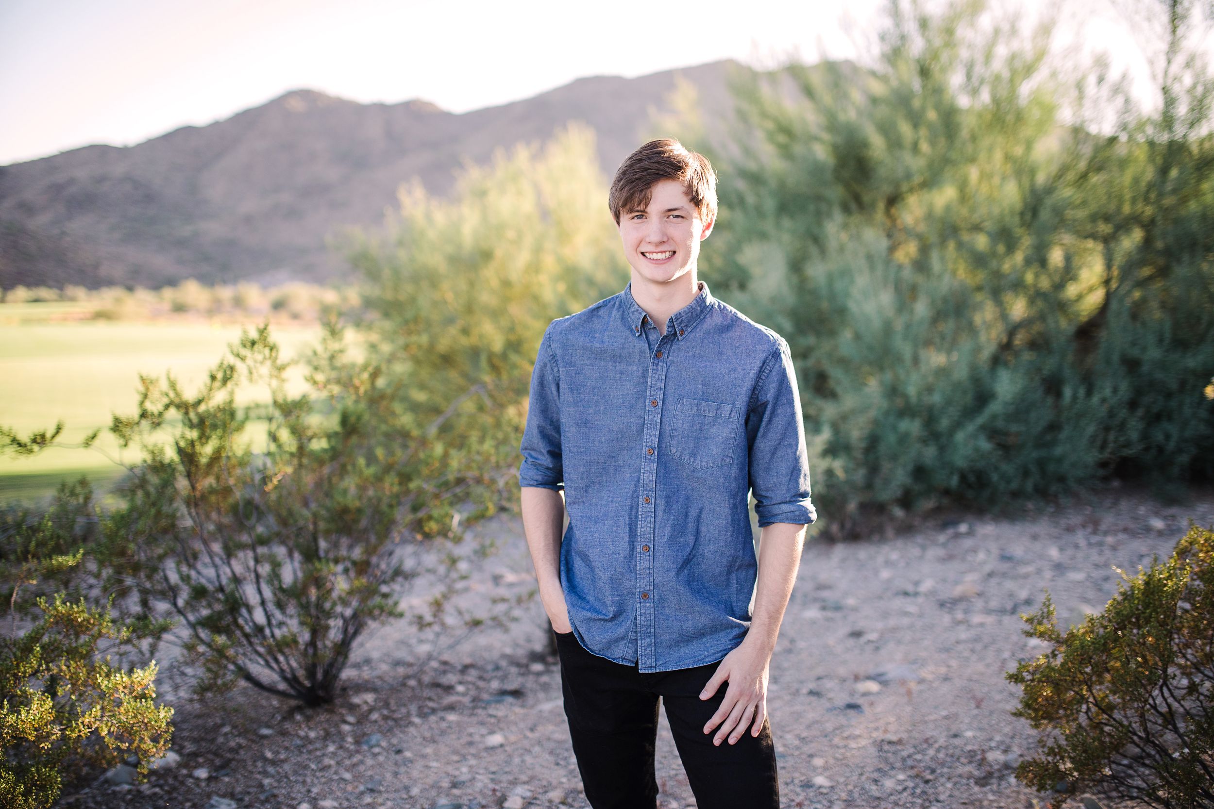 Smiling young man in blue denim shirt standing in desert landscape with mountains.