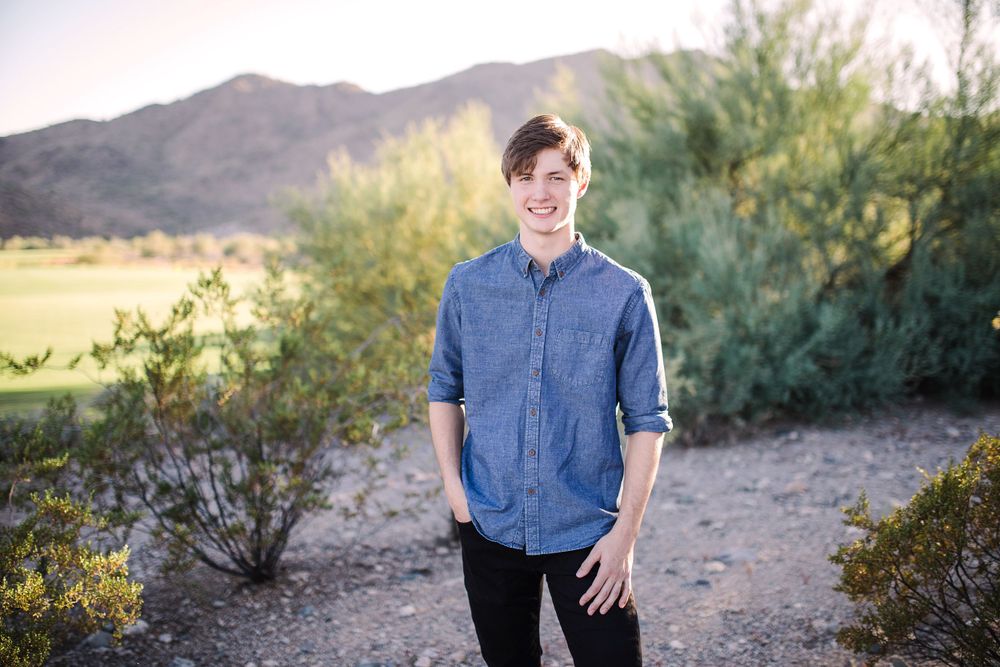 Smiling young man in blue denim shirt standing in desert landscape with mountains.