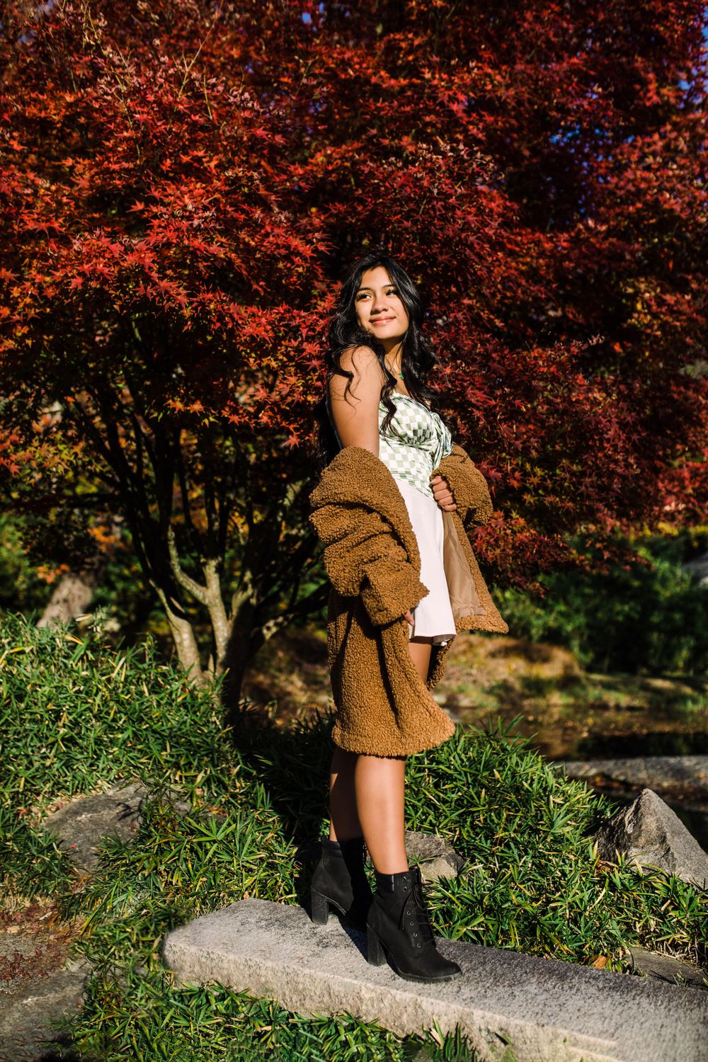 Young woman in white dress and brown teddy coat poses before striking red Japanese maple tree.