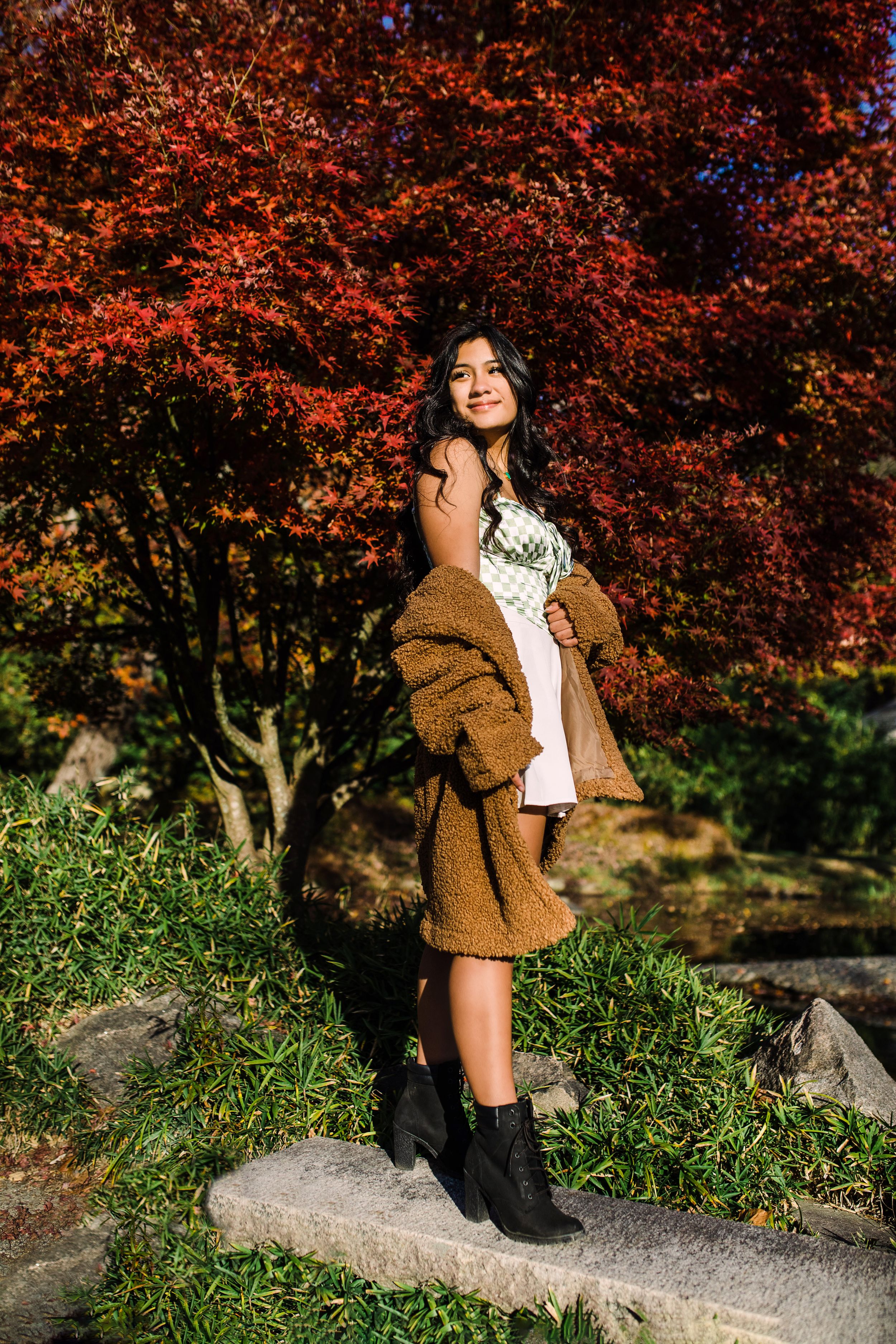 Young woman in white dress and brown teddy coat poses before striking red Japanese maple tree.