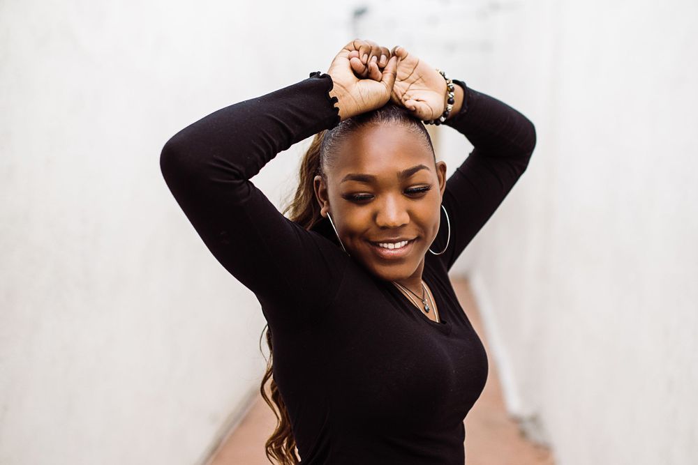 Young Black woman in black long-sleeve top smiles while styling her hair against a white wall.