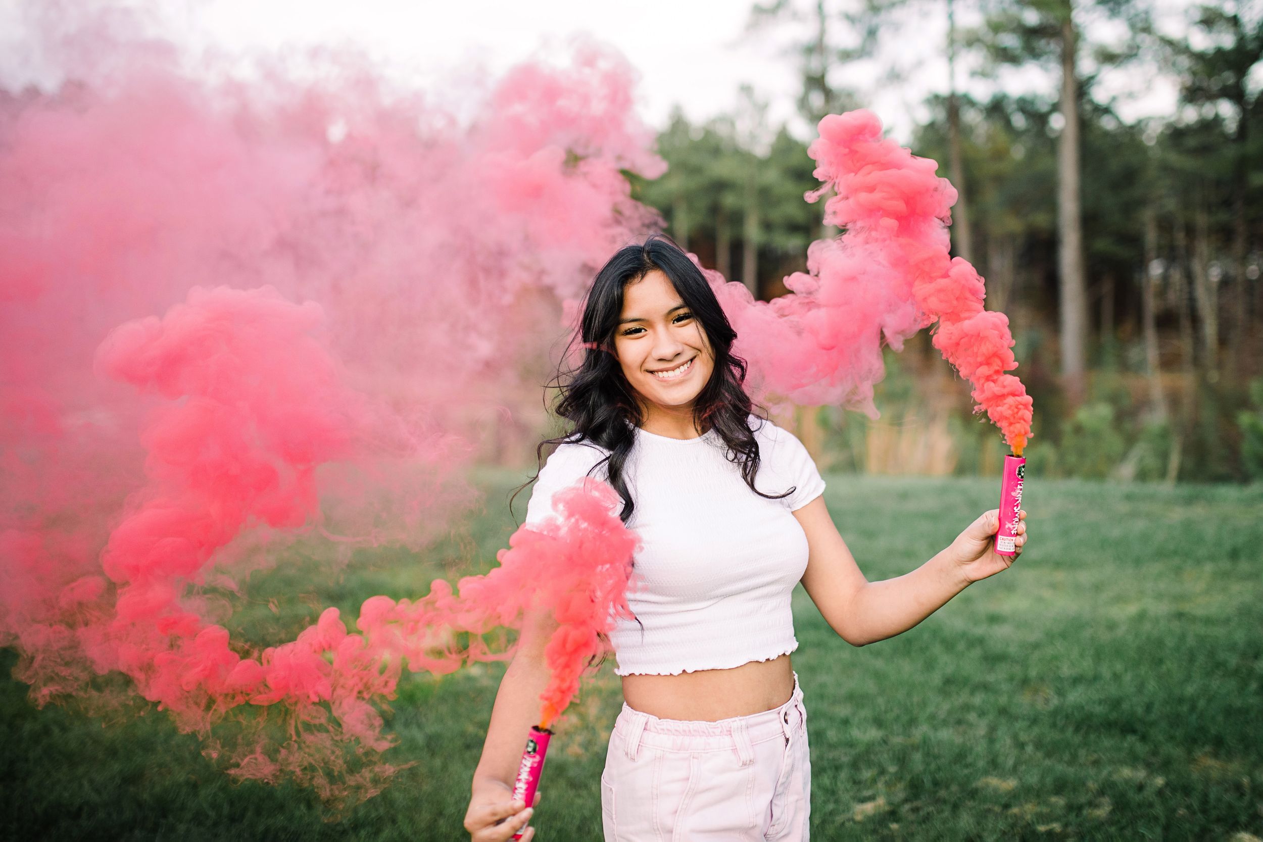 Young woman in white crop top smiling while holding pink smoke bombs in a green field.