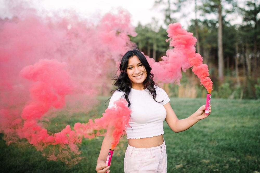 Young woman in white crop top smiling while holding pink smoke bombs in a green field.
