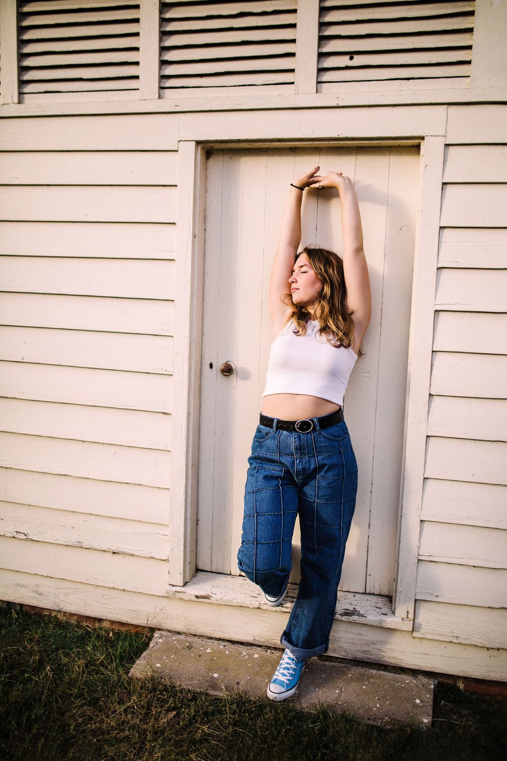 Young woman in white crop top and jeans posing with arms raised in a white wooden doorway.