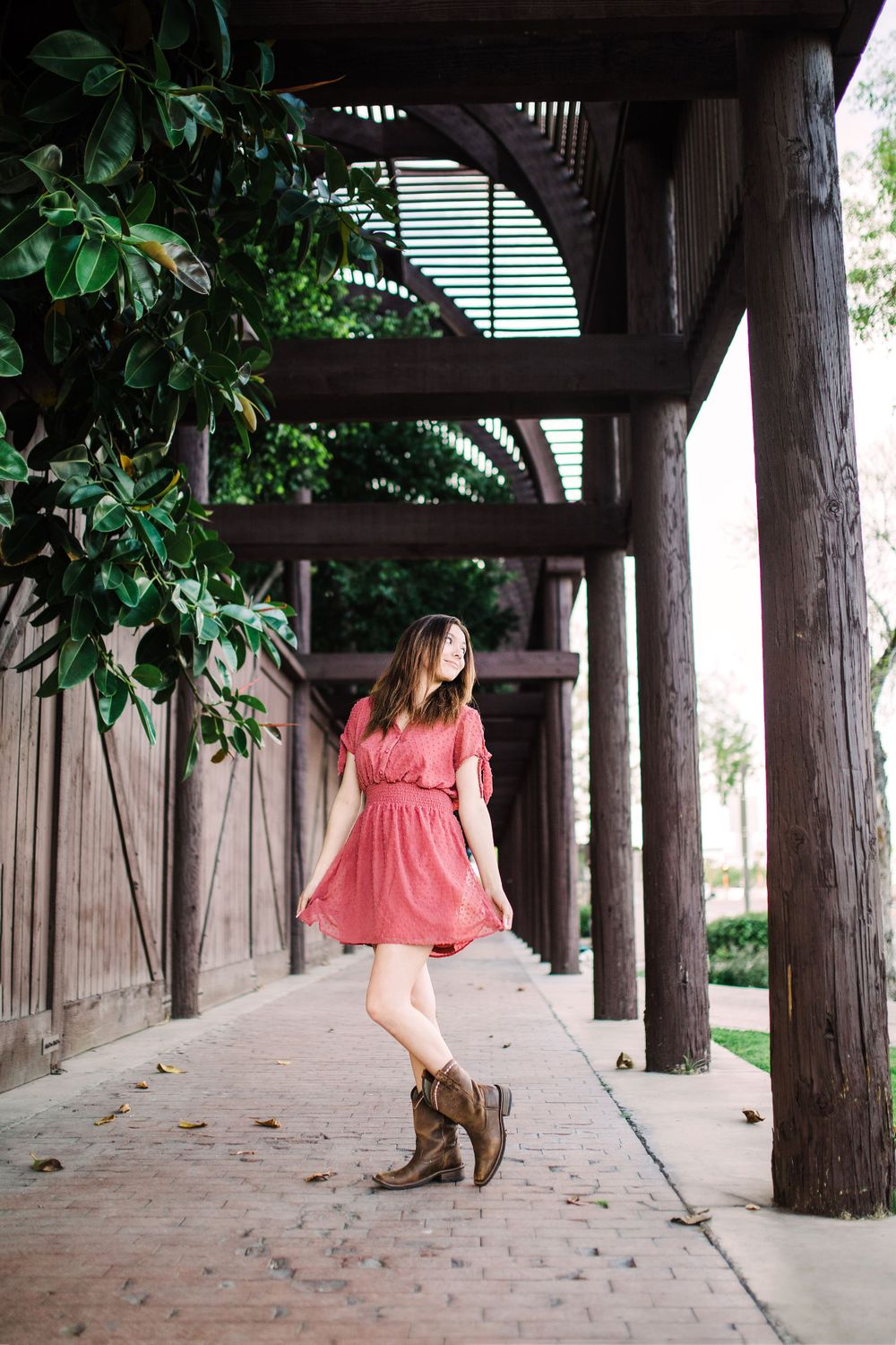 Young woman in pink dress and brown boots walking under wooden pergola with lush green foliage.