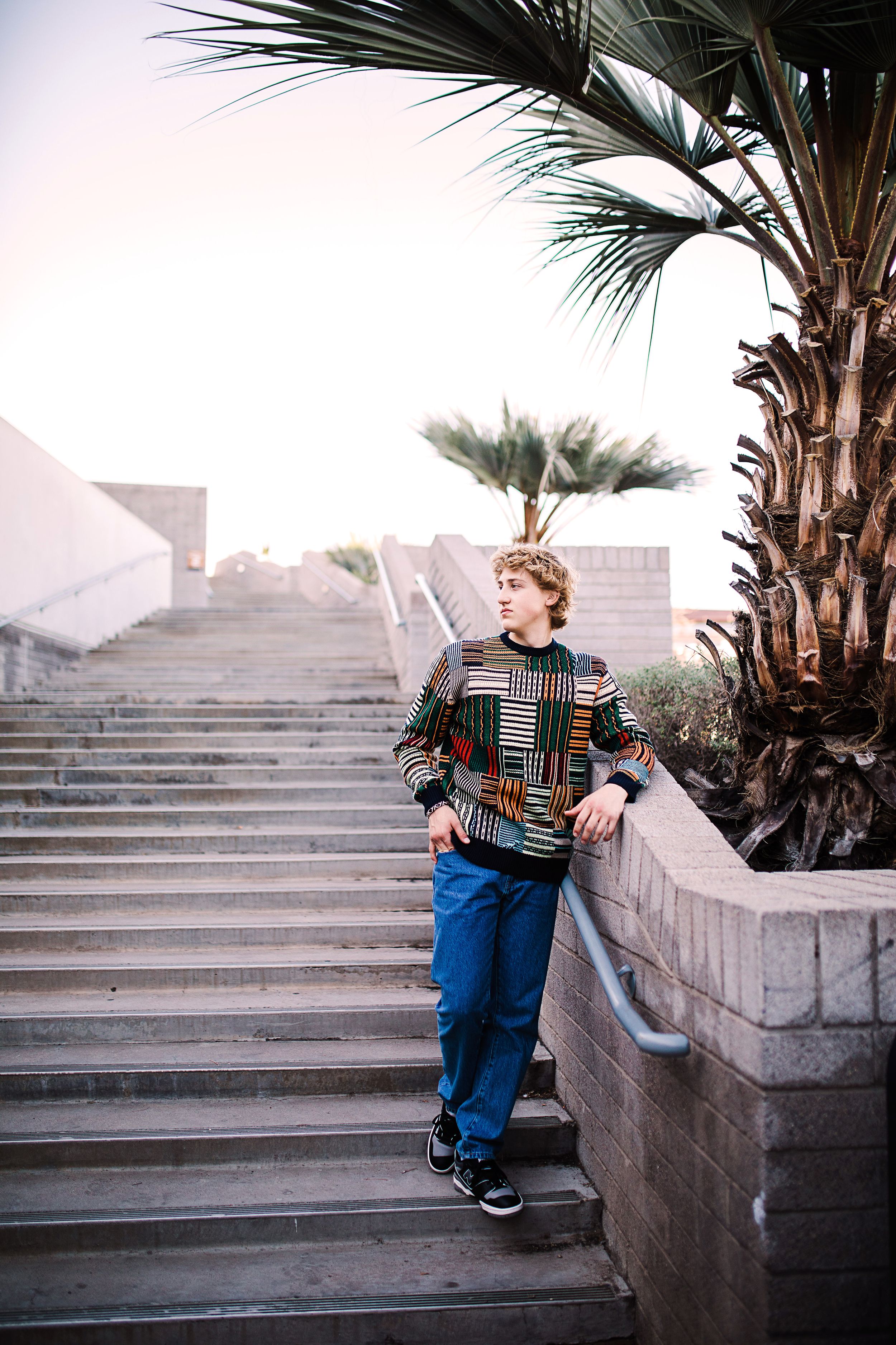 Young man in colorful patterned sweater and jeans leaning on stair railing near palm tree outdoors.