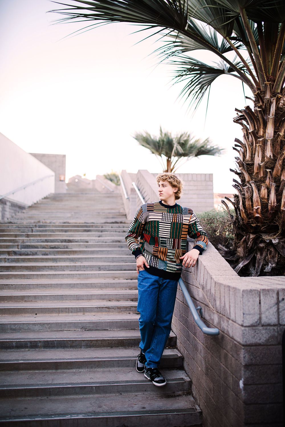 Young man in colorful patterned sweater and jeans leaning on stair railing near palm tree outdoors.