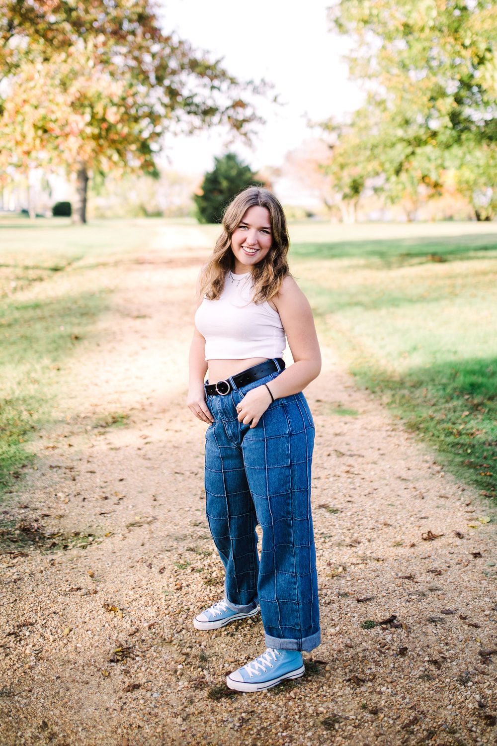 Smiling young woman in white crop top and wide-leg jeans posing on a dirt path in autumn park.