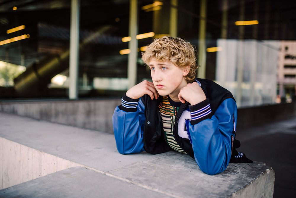 Young teen boy in blue varsity jacket resting on concrete ledge looking pensive.
