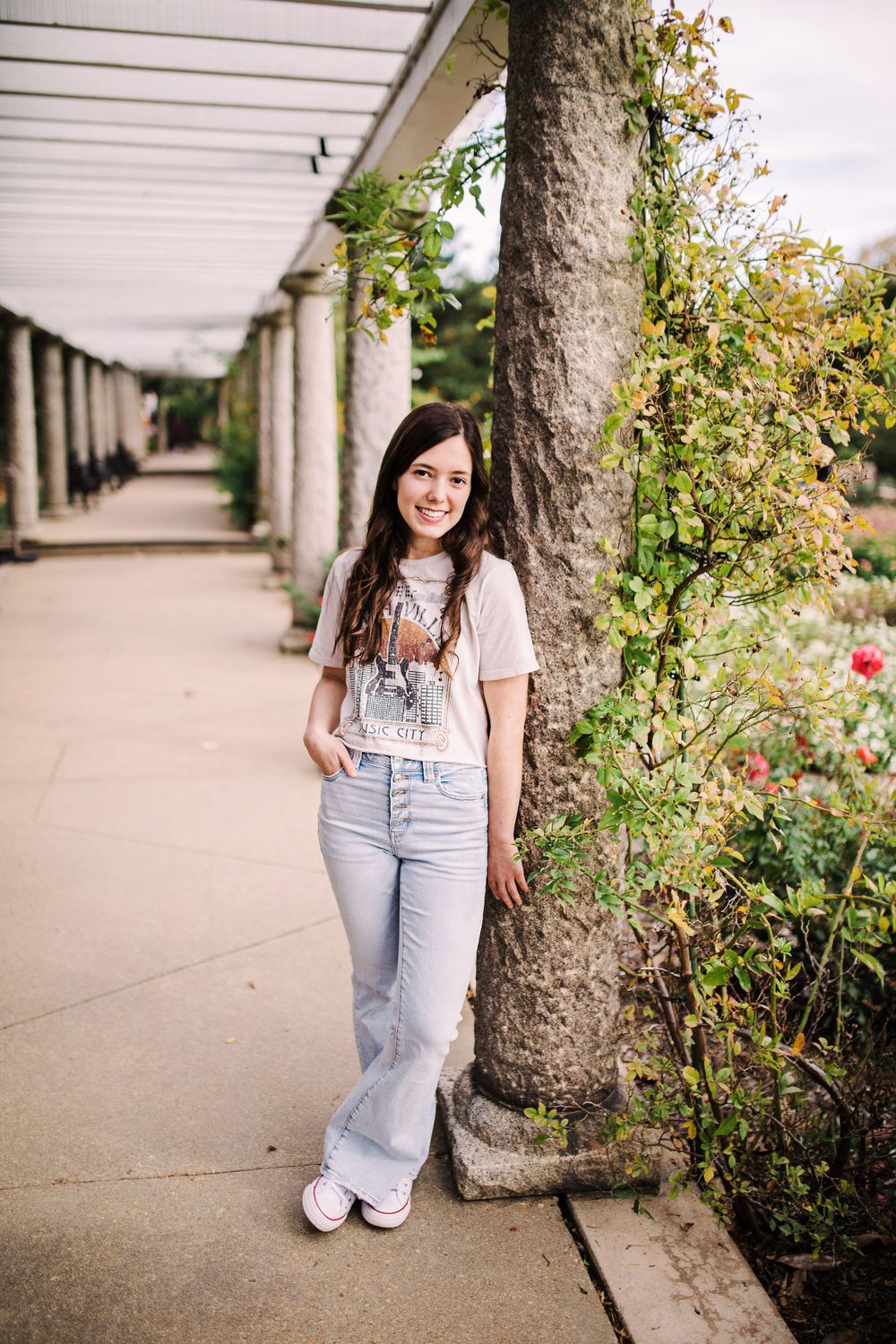 Smiling young woman in graphic tee and flare jeans leaning against ivy-covered column in pergola.