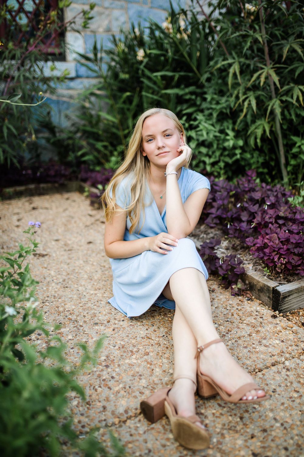Blonde teen girl in light blue dress sitting in garden with purple flowers and green foliage.