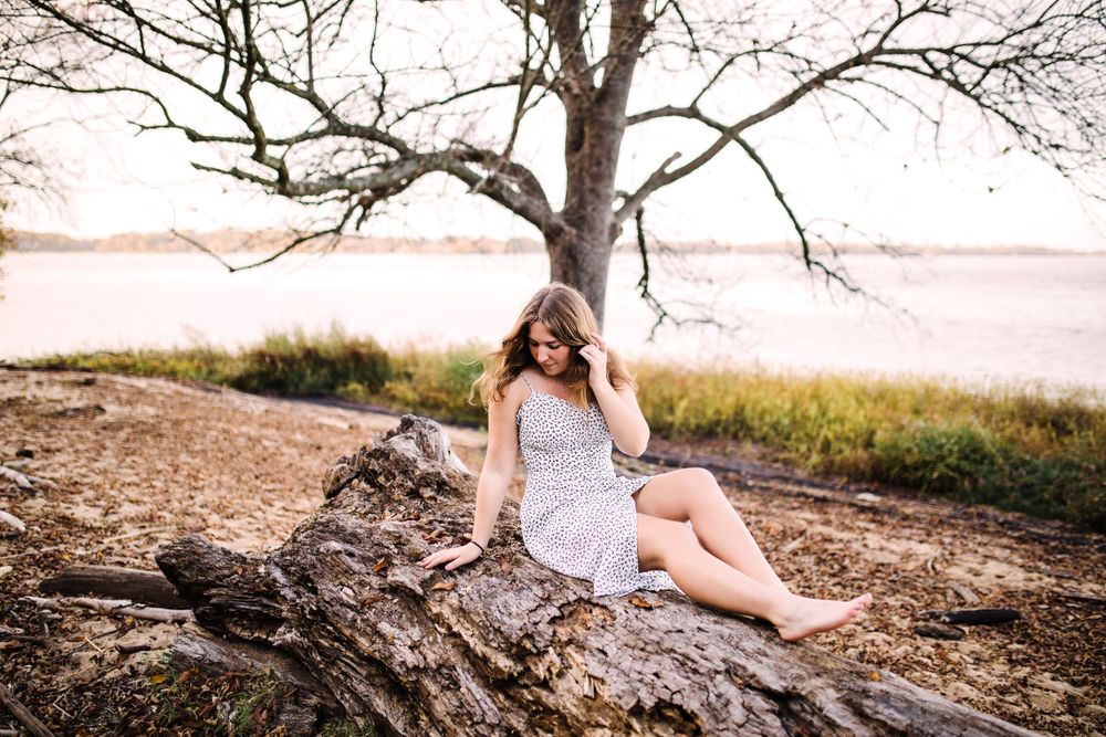 Young woman in floral dress sitting on fallen log by lake under bare tree at golden hour.