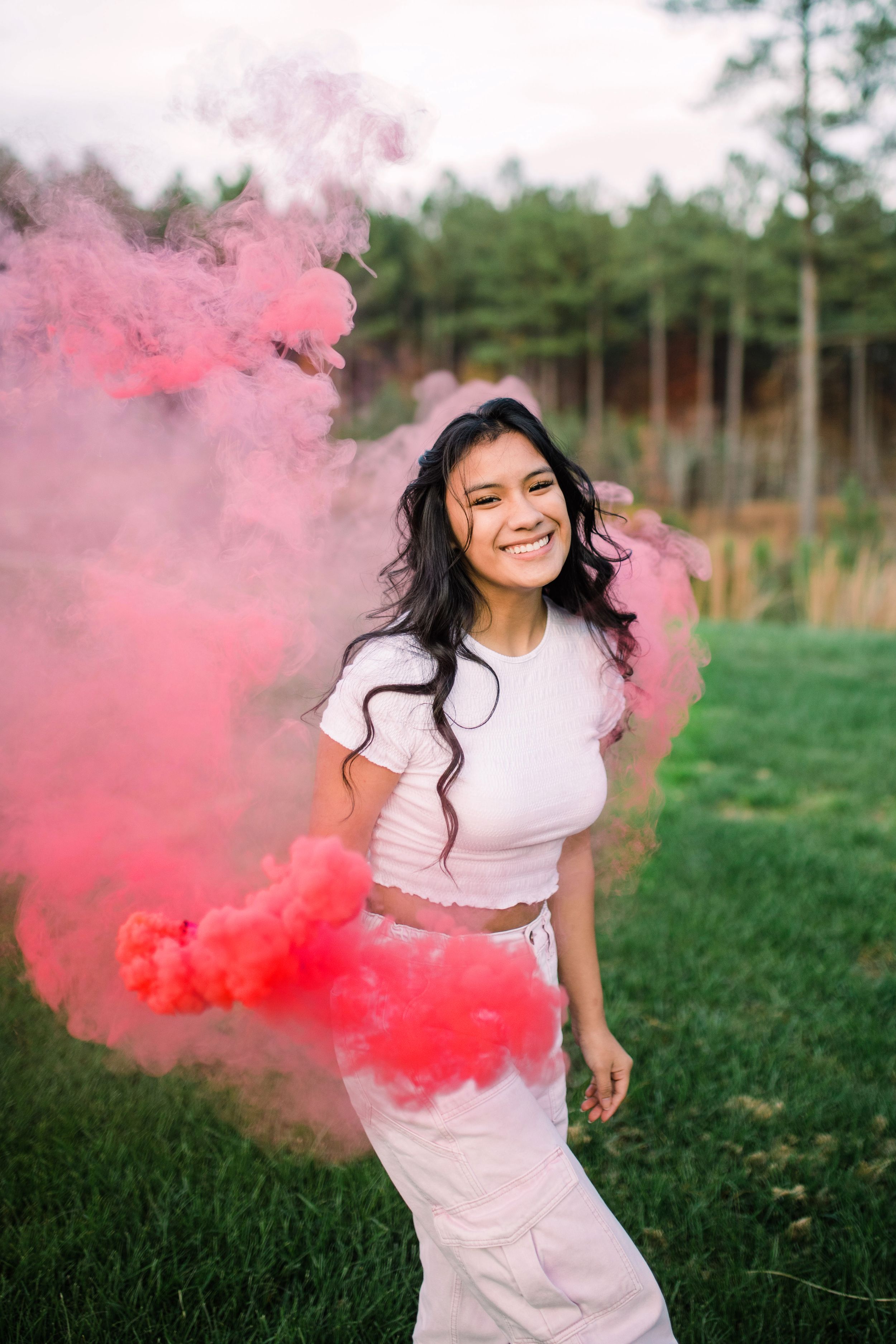 Young woman in white outfit laughing joyfully surrounded by billowing pink and red smoke outdoors.