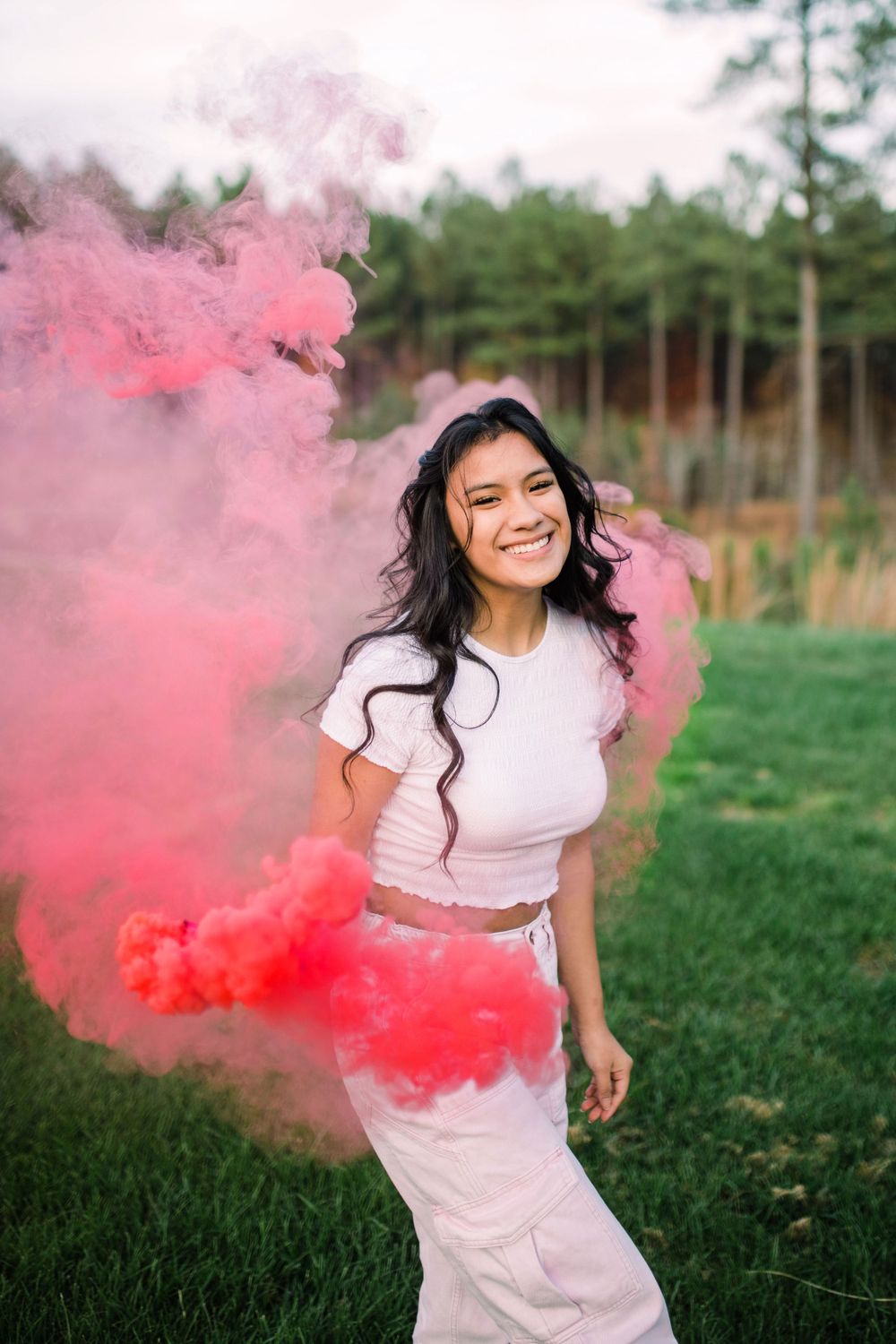 Young woman in white outfit laughing joyfully surrounded by billowing pink and red smoke outdoors.