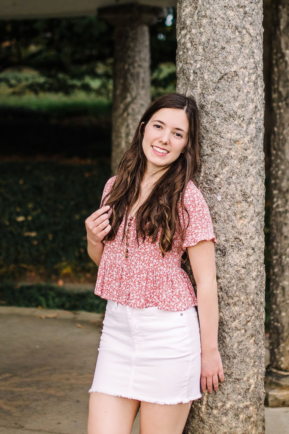 Smiling young woman in pink floral top and white mini skirt leaning against stone column in garden.