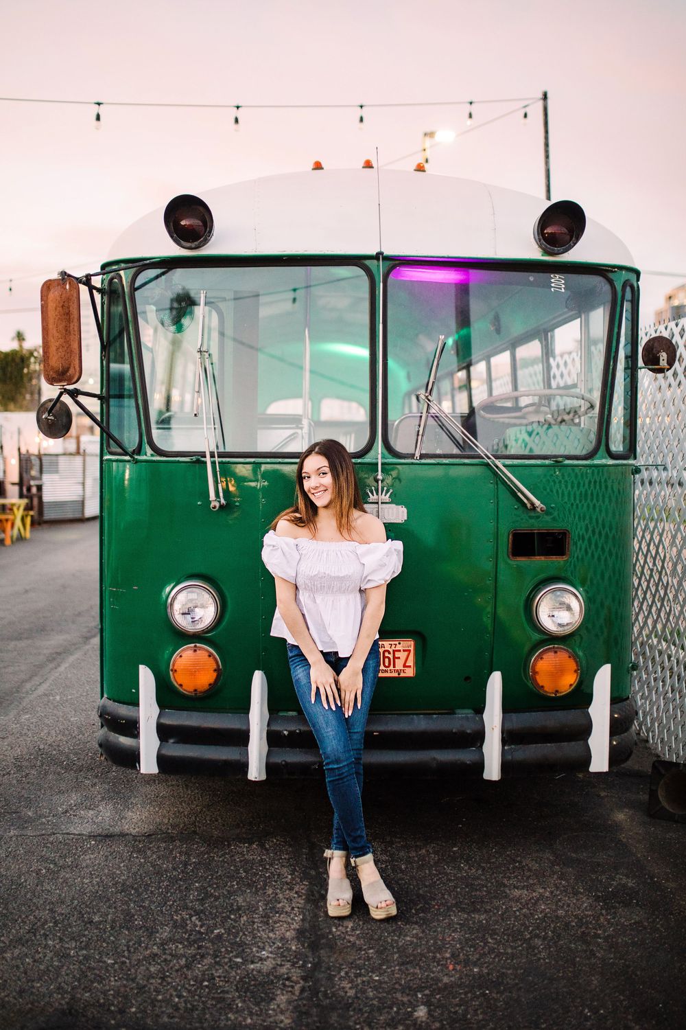 Young woman in white top and jeans sitting on vintage green bus at outdoor venue at dusk.