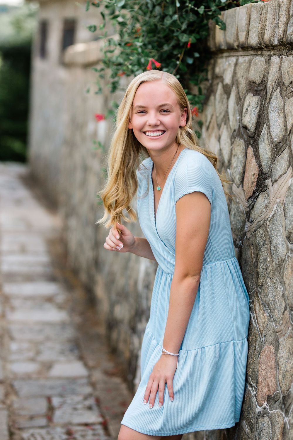 Smiling blonde teenage girl in light blue dress leans against stone wall in garden pathway.