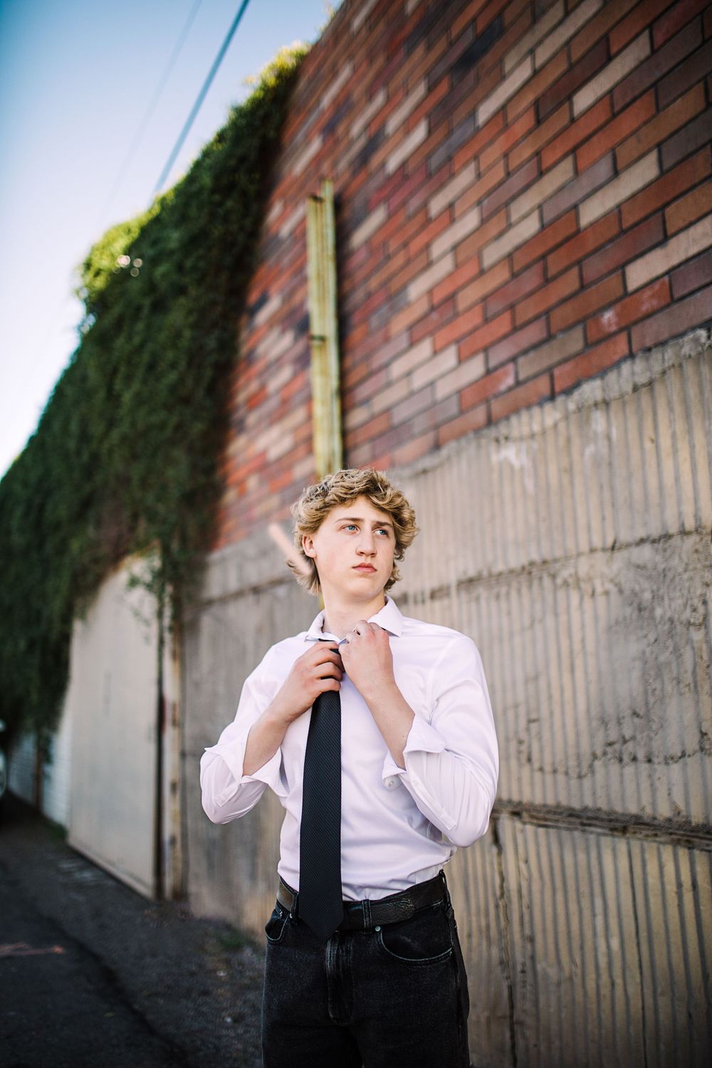 Young man in white shirt adjusting black tie standing in alley with ivy-covered brick wall.