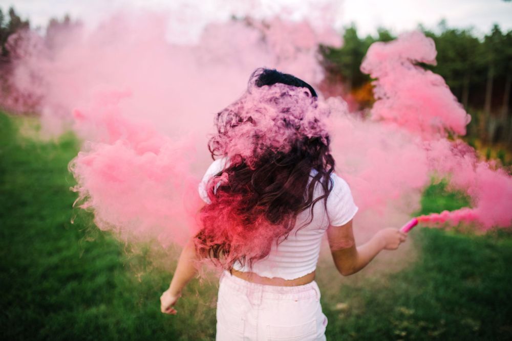 Girl with dark hair swinging pink smoke bomb outdoors in green field.