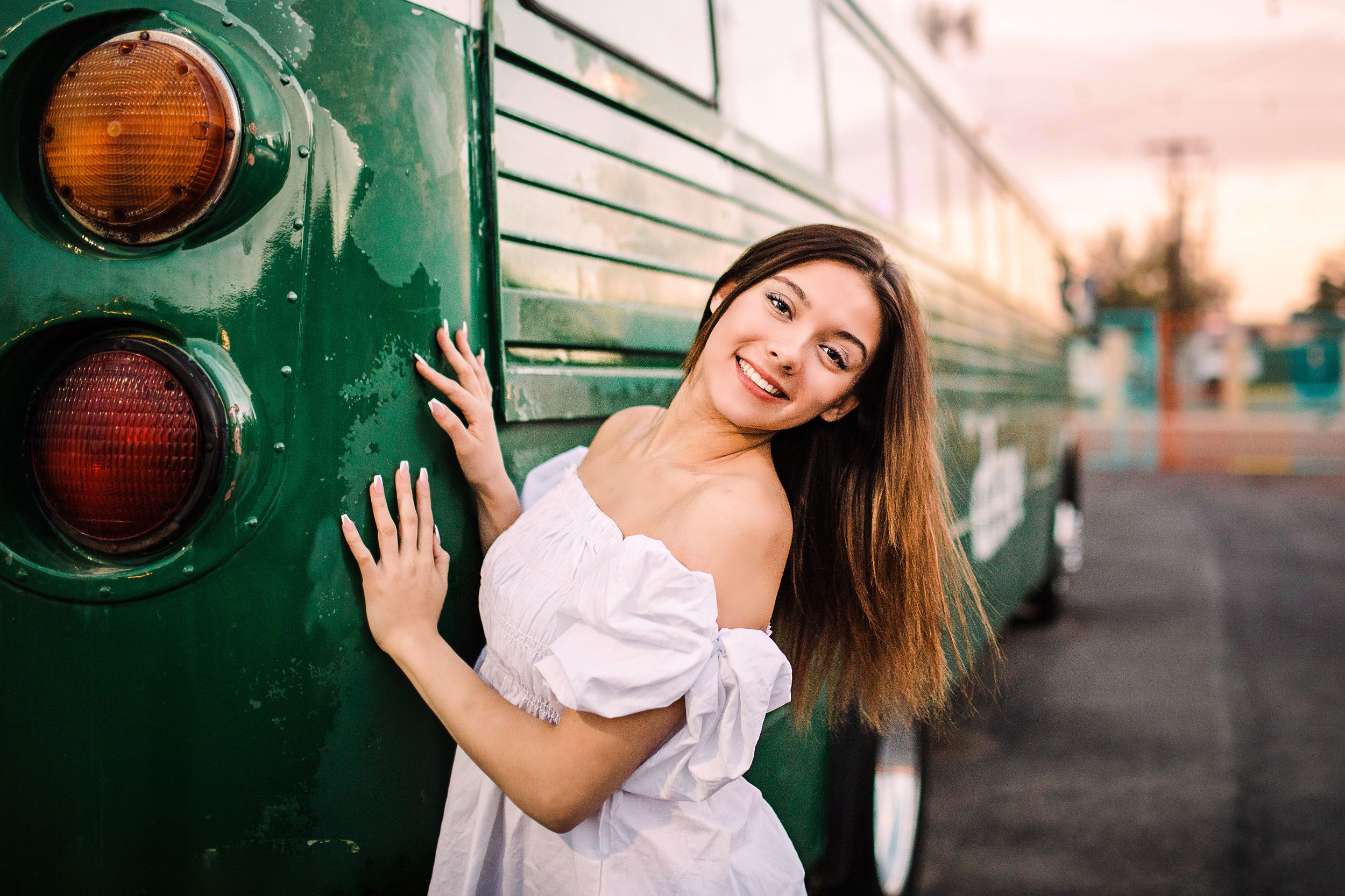 Young woman in white off-shoulder dress leaning against vintage green bus at sunset.