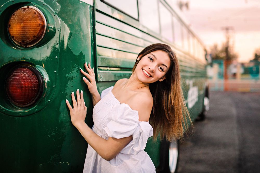 Young woman in white off-shoulder dress leaning against vintage green bus at sunset.