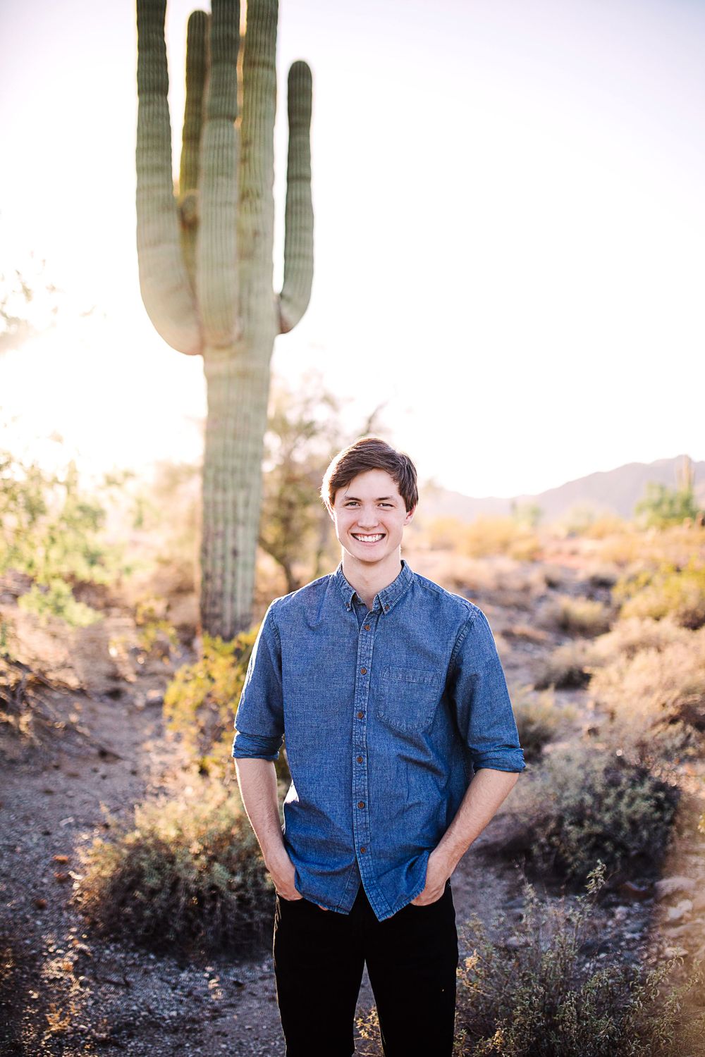 Smiling teen boy in denim shirt standing before tall saguaro cactus in Arizona desert.
