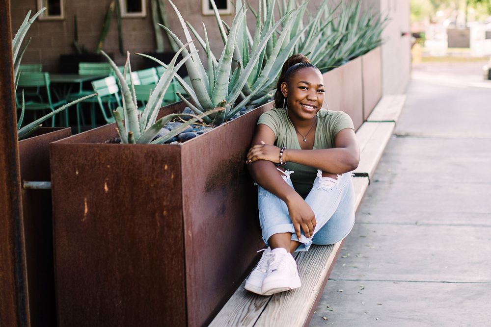 Laughing young Black woman in olive tee and jeans sits on sidewalk beside rustic metal planter with agave.