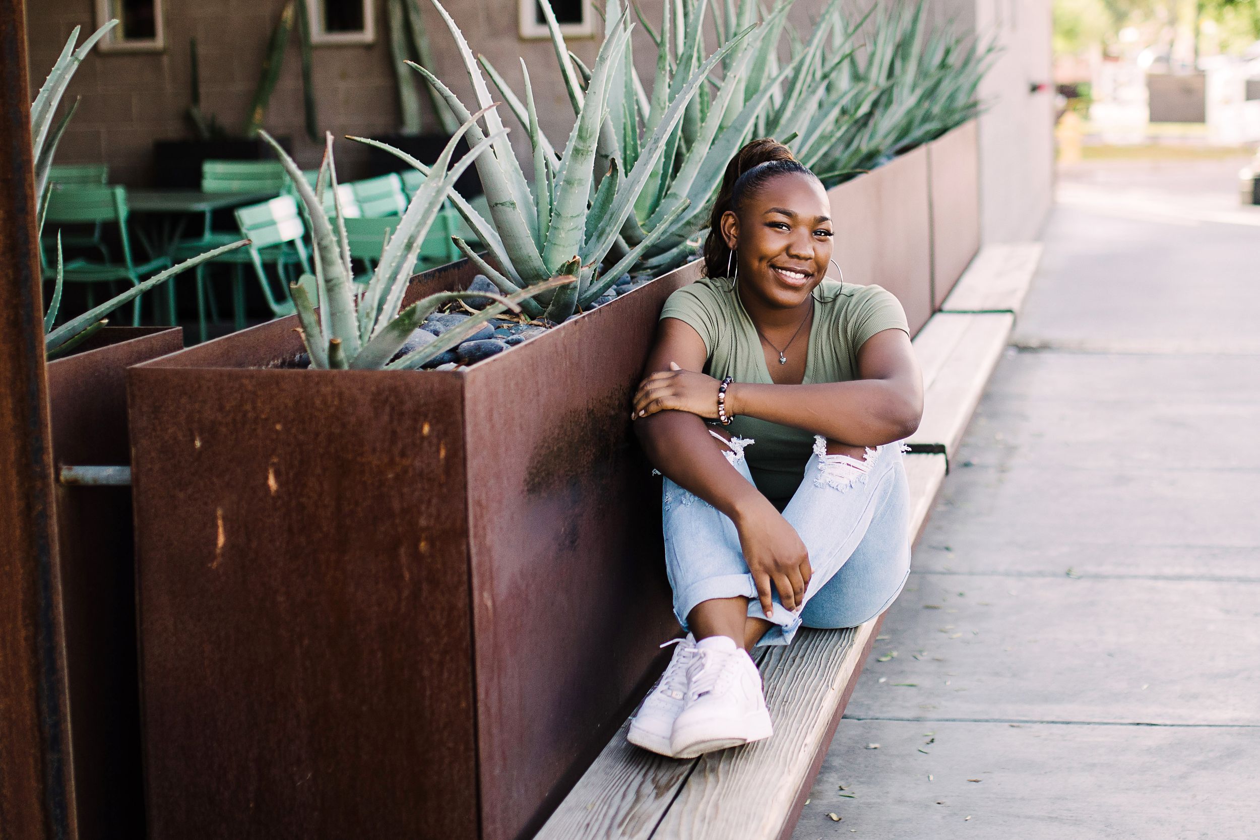 Laughing young Black woman in olive tee and jeans sits on sidewalk beside rustic metal planter with agave.