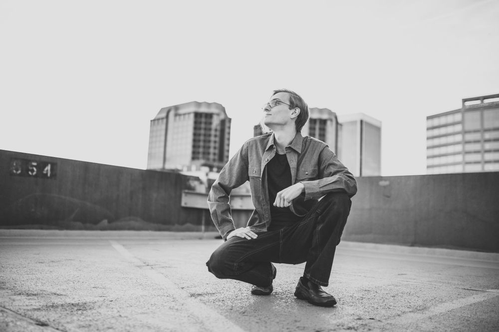 Black and white photo of person crouching on urban rooftop with city buildings in background.