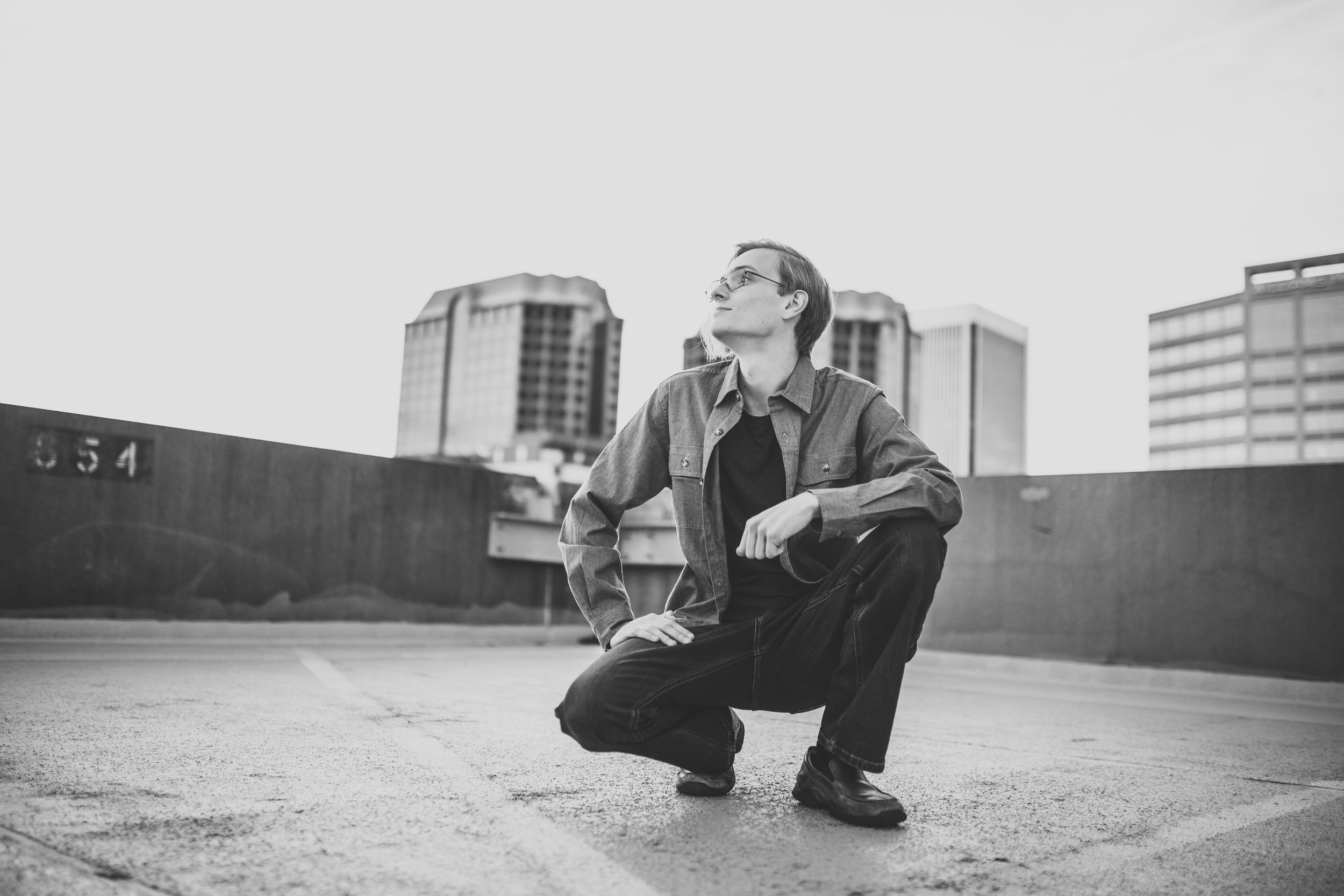 Black and white photo of person crouching on urban rooftop with city buildings in background.