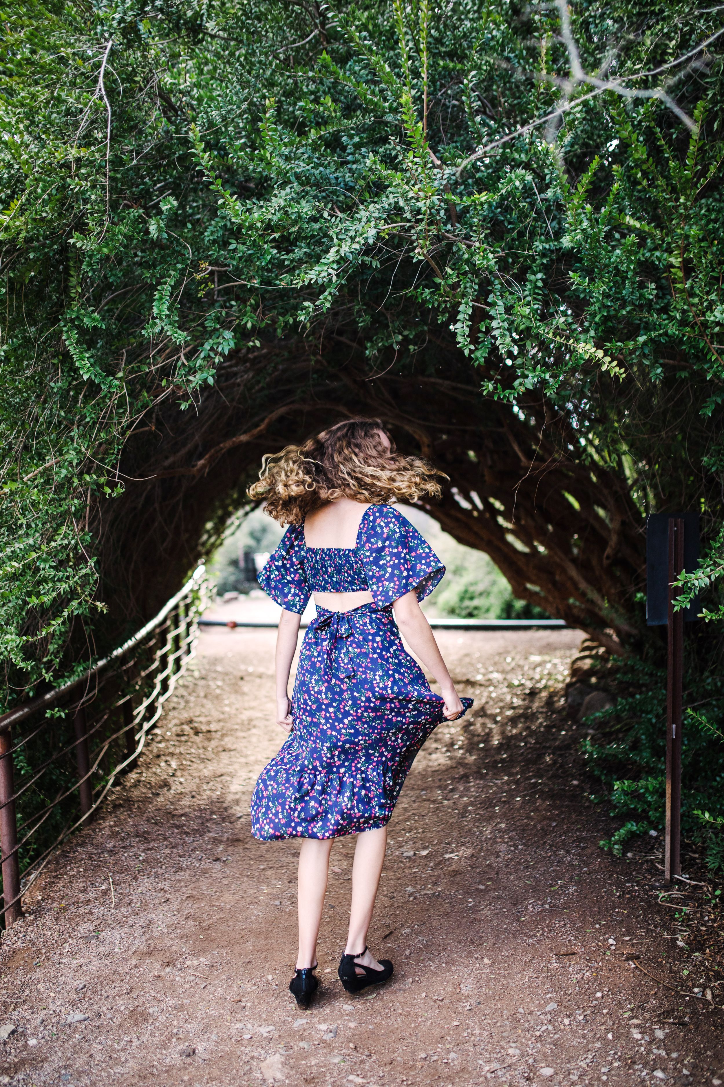 Woman in blue floral dress walking through a natural green archway of trees on a dirt path.