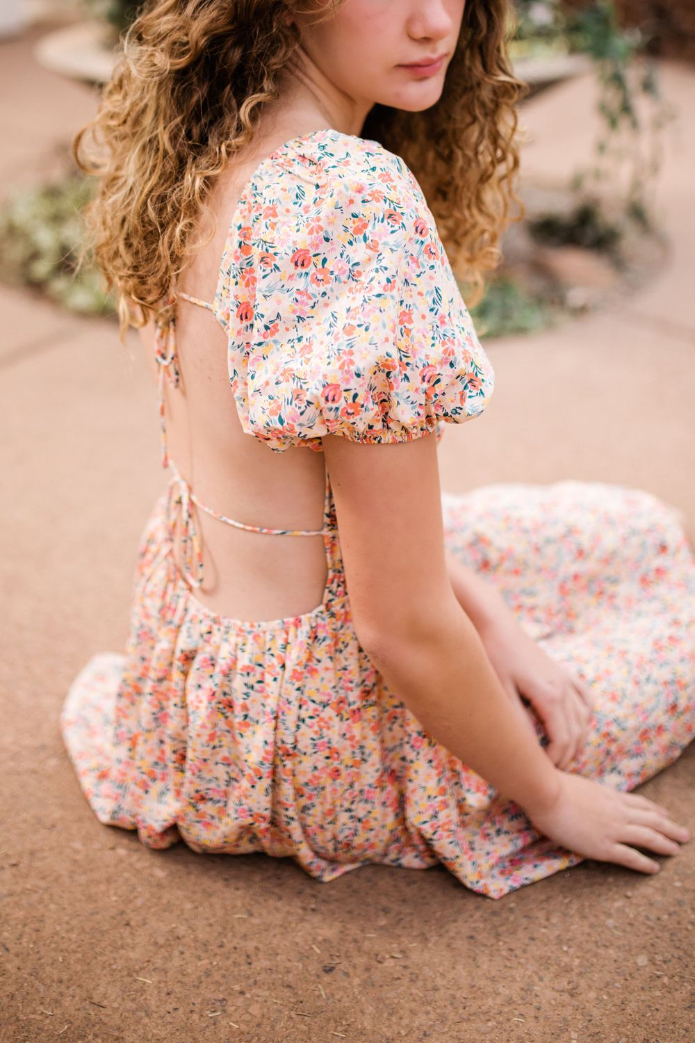 Back view of young woman in open-back floral dress sitting outdoors near plants.