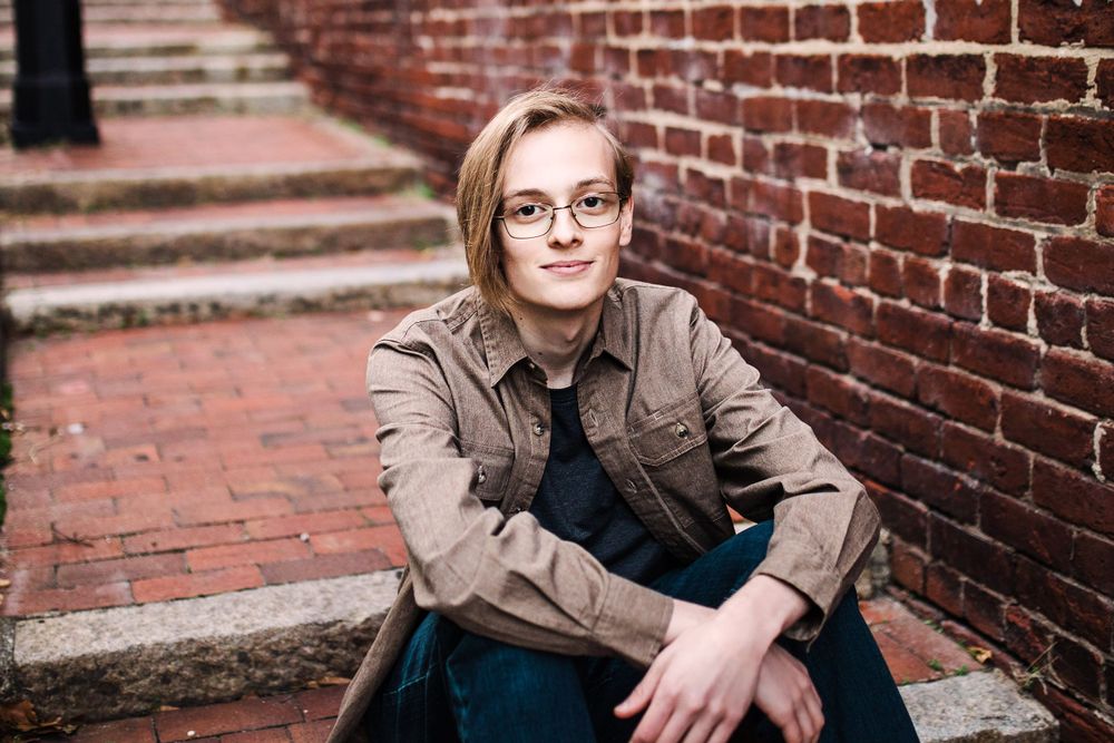 Young person with glasses sitting on brick steps against red brick wall in urban setting.