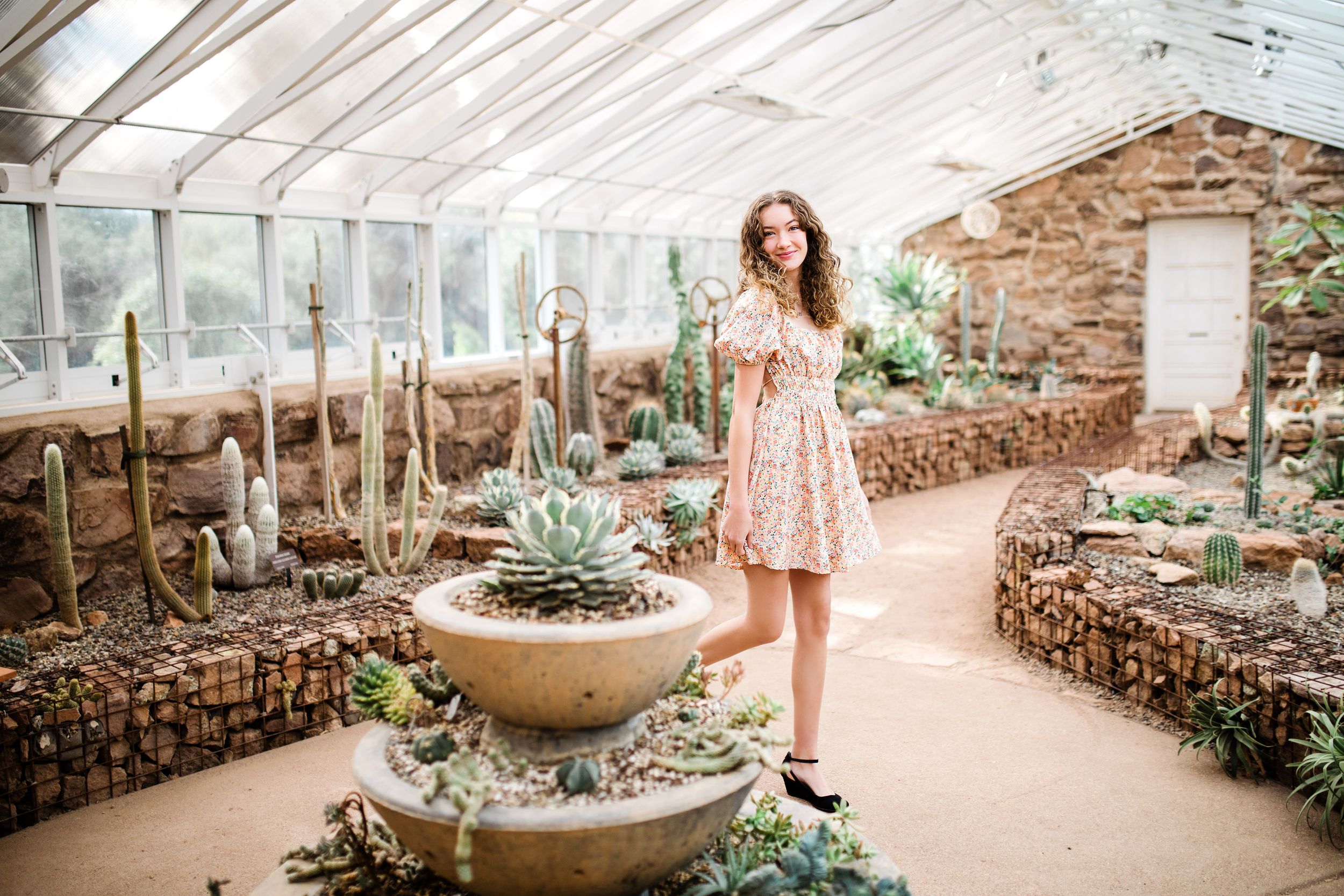 Young woman in pink floral dress standing near large succulent display inside glass greenhouse.