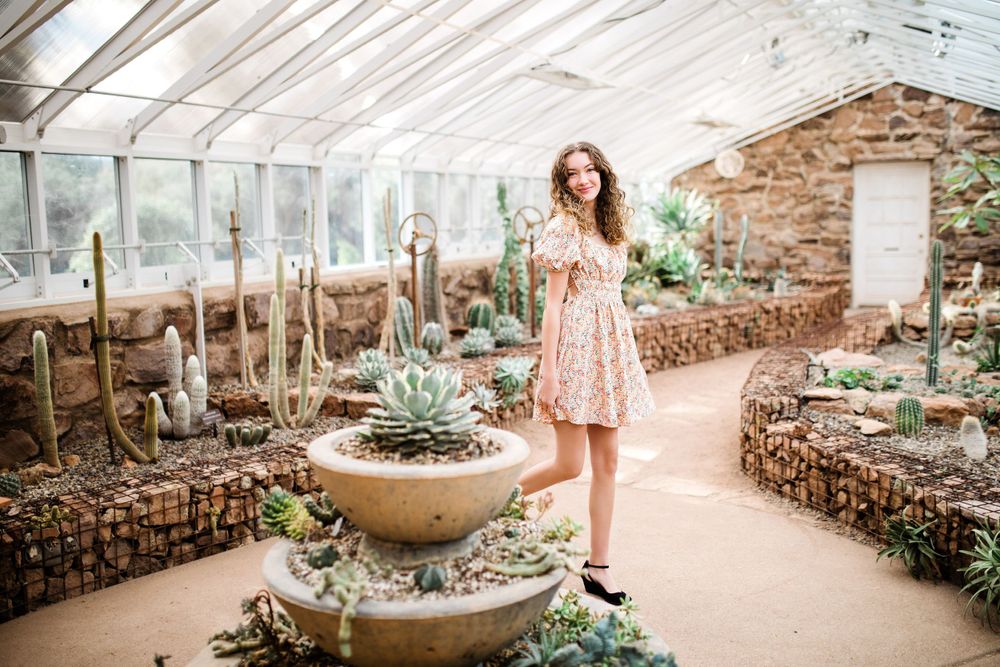 Young woman in pink floral dress standing near large succulent display inside glass greenhouse.