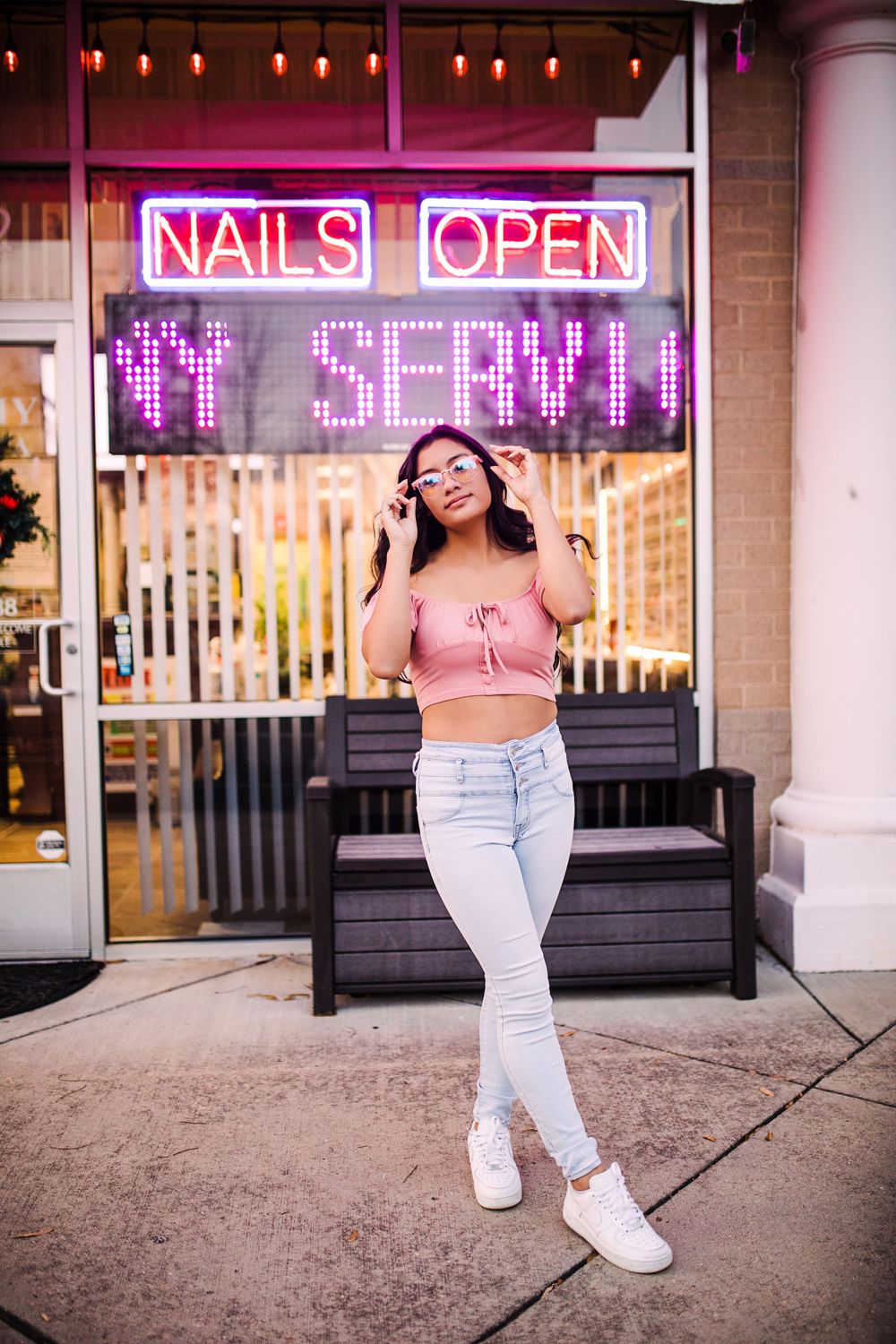 Young woman in pink crop top and jeans posing in front of neon nail salon open sign.