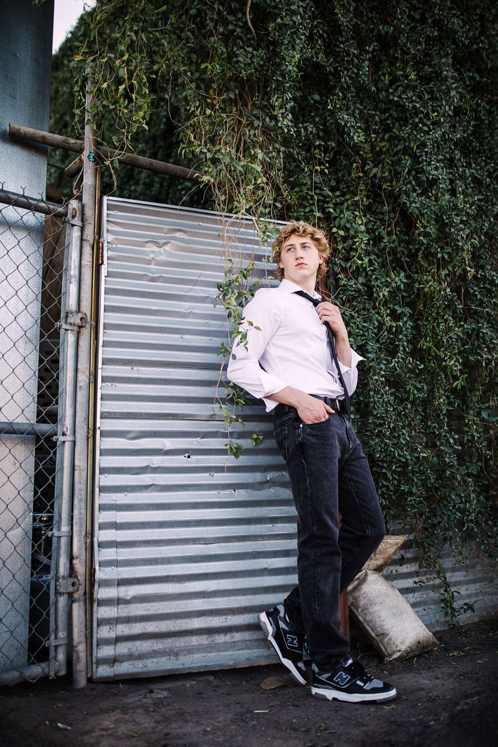 Young man in white shirt and black tie leaning on corrugated metal gate with ivy-covered wall.