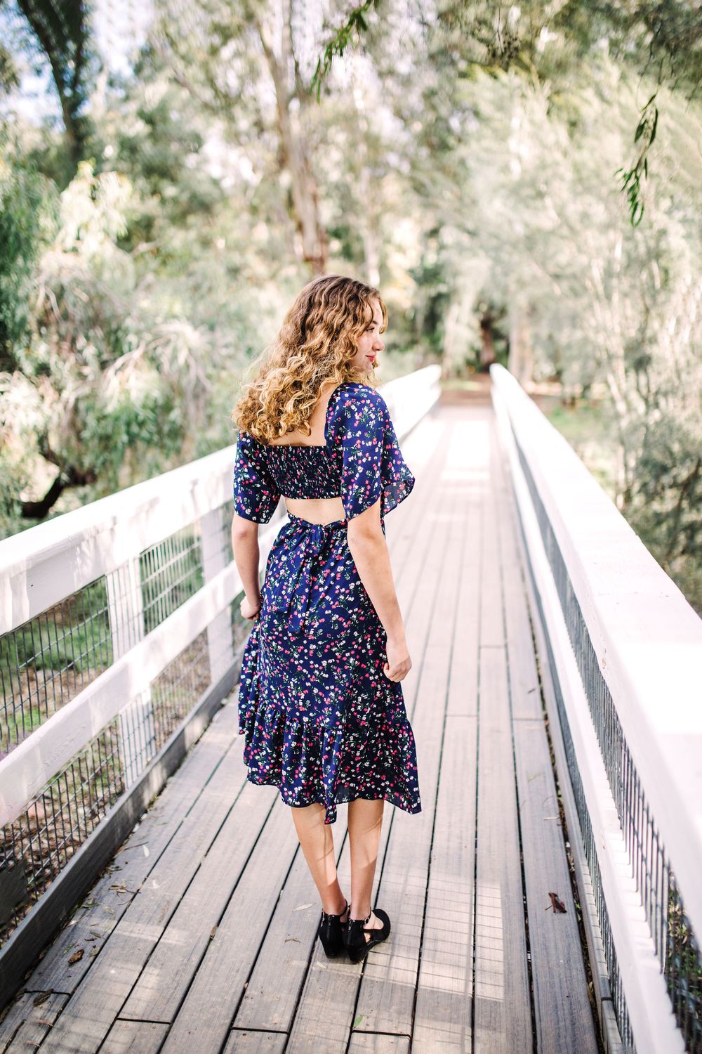 Young woman in navy floral midi dress with cutout back walking on a wooden bridge.