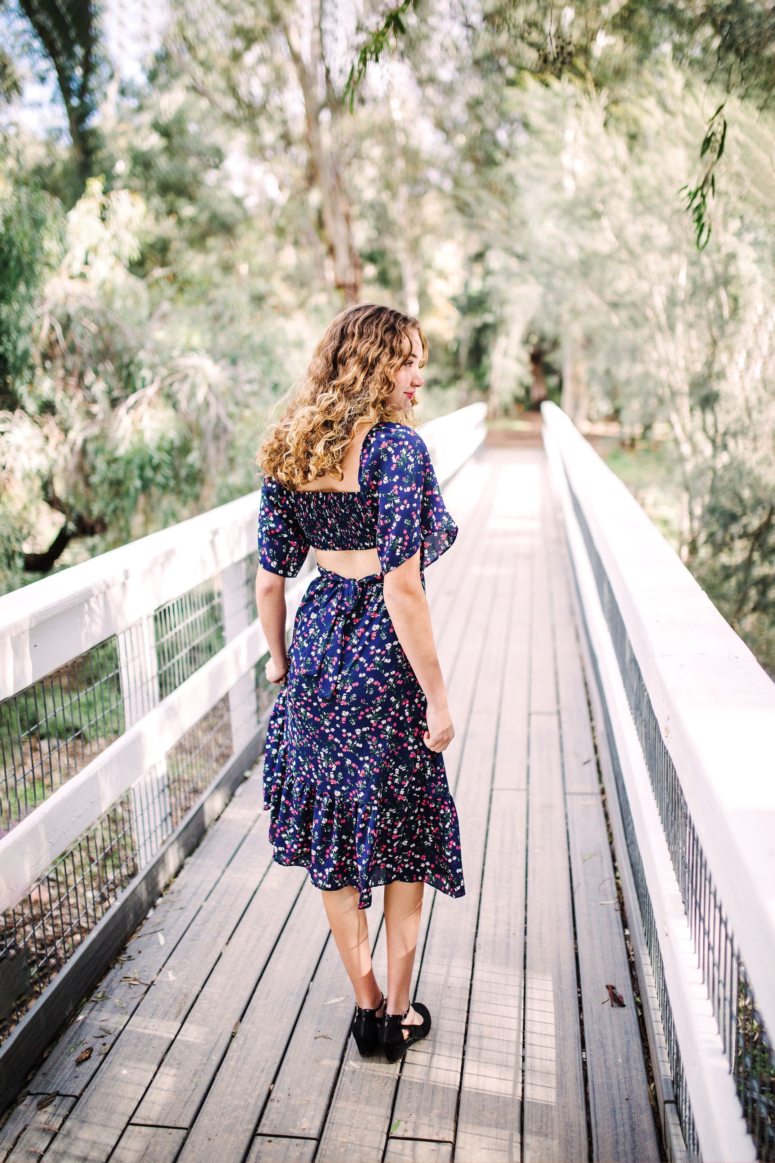 Young woman in navy floral midi dress with cutout back walking on a wooden bridge.