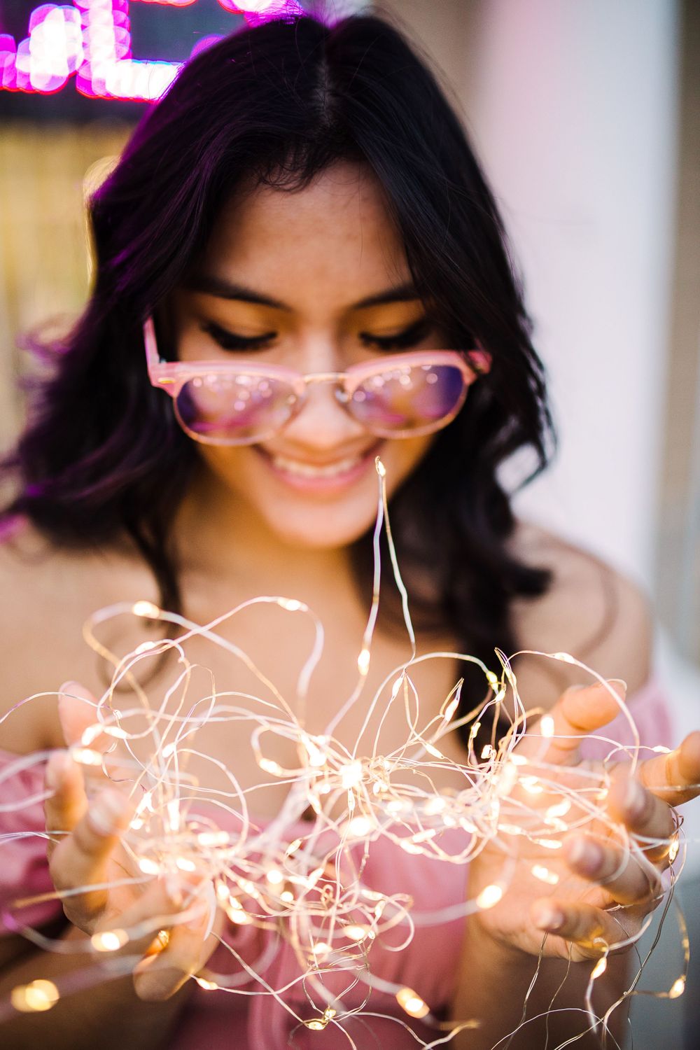 Smiling young woman holding glowing fairy lights with pink sunglasses pushed down her nose.