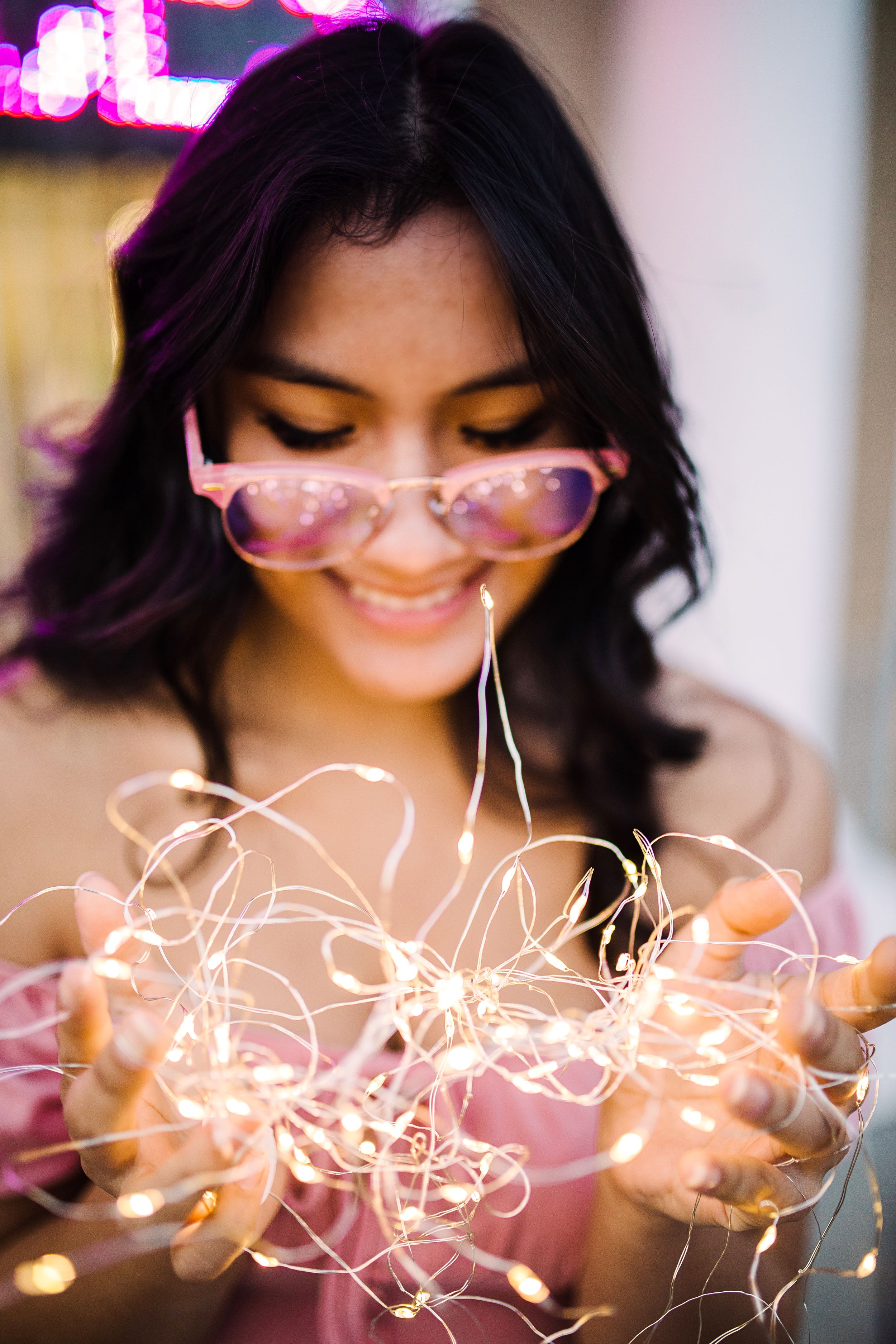 Smiling young woman holding glowing fairy lights with pink sunglasses pushed down her nose.