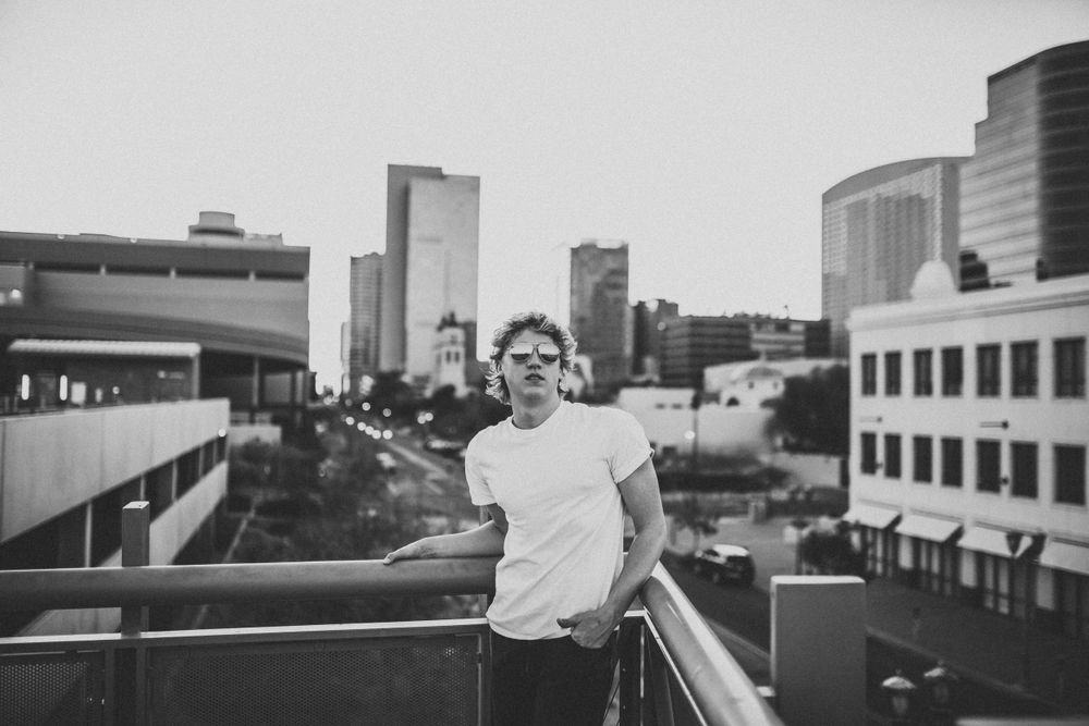 Young man leaning on railing with city skyline in black and white urban photo.