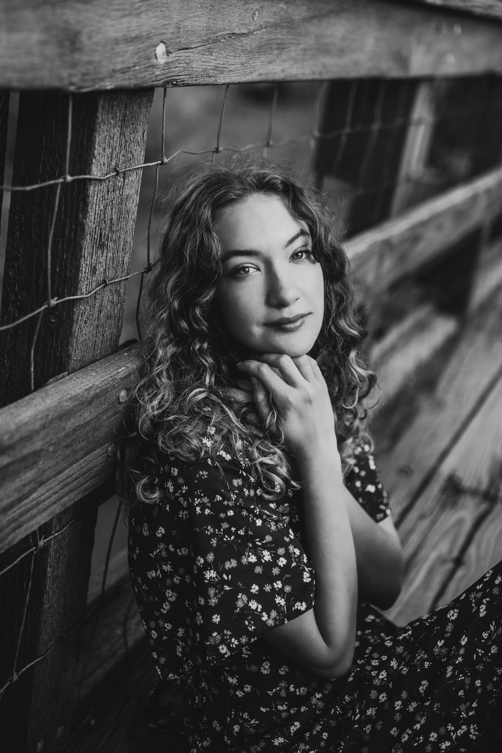 Black and white portrait of a curly-haired young woman in floral dress leaning against a wooden fence.