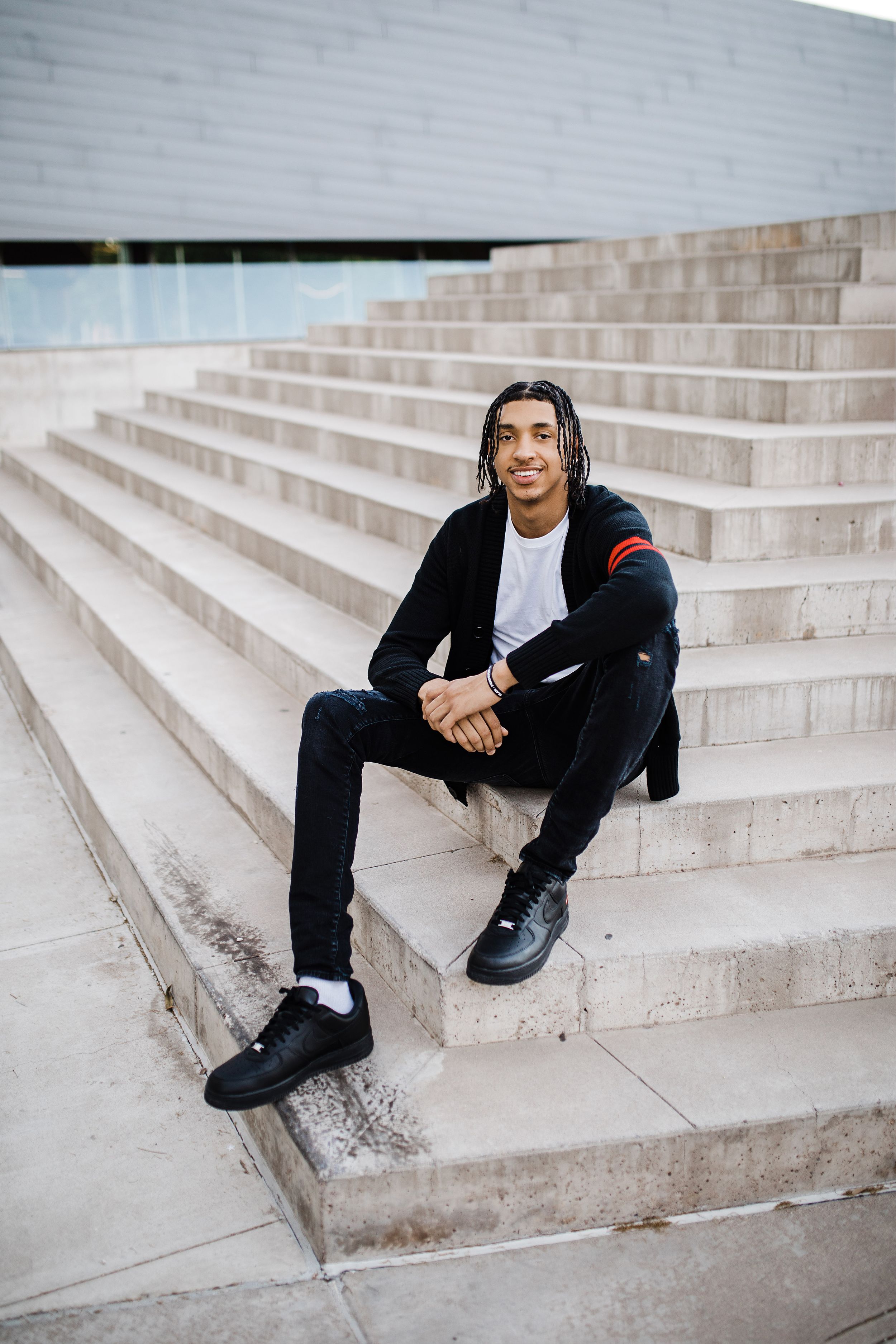 Smiling young man with braids in black outfit sits on concrete steps of modern building.