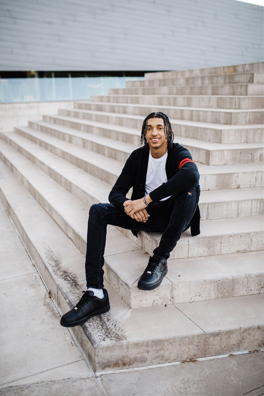 Smiling young man with braids in black outfit sits on concrete steps of modern building.