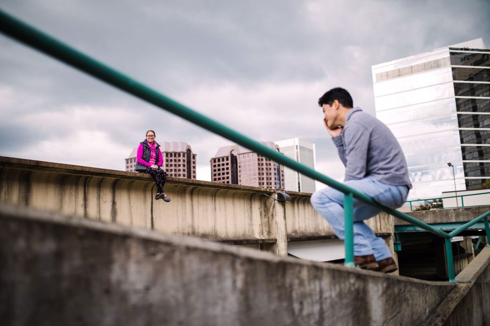 Young man leaning on concrete railing with woman sitting in background on urban rooftop.