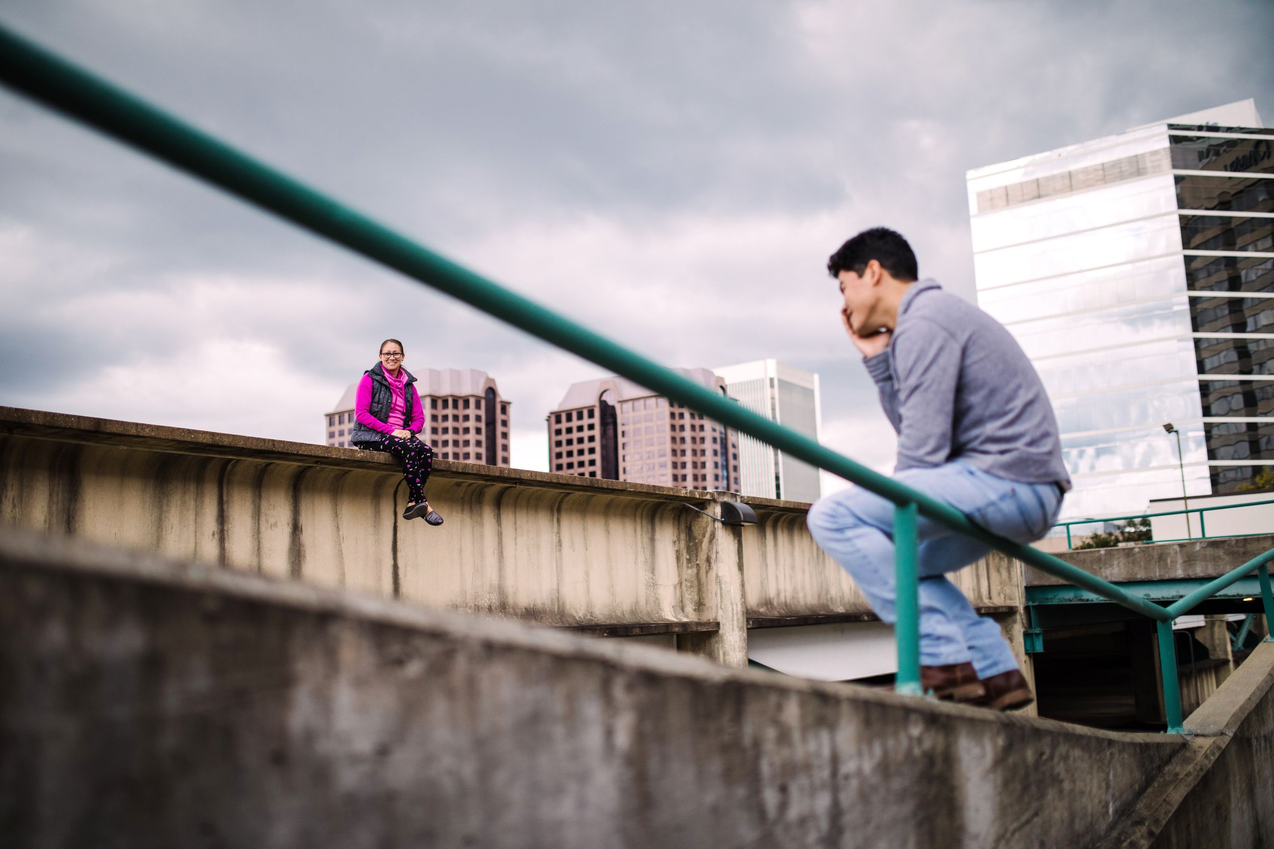 Young man leaning on concrete railing with woman sitting in background on urban rooftop.