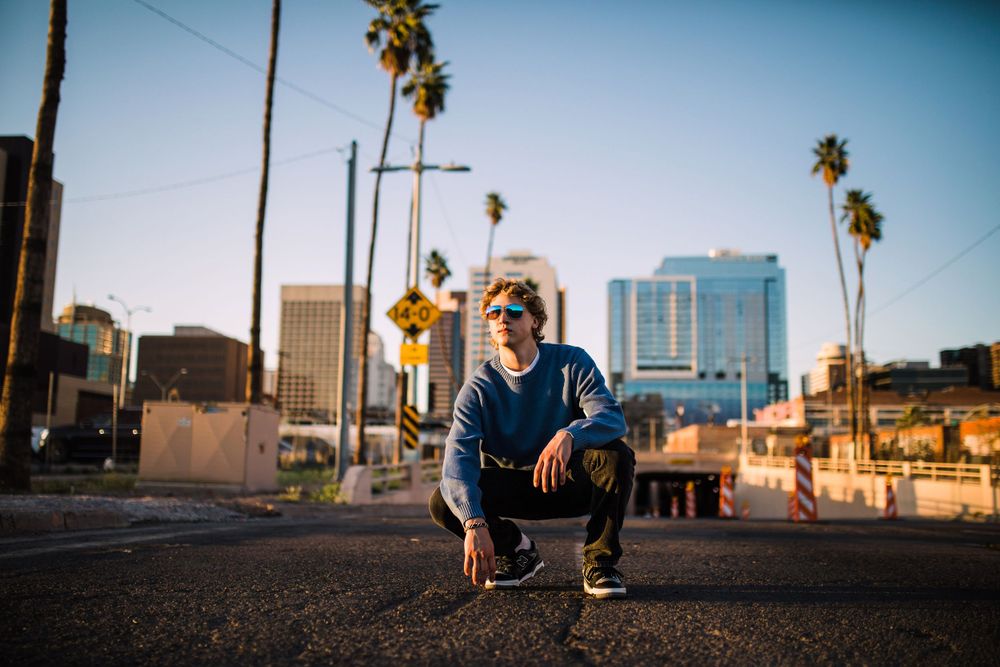 Young man crouching on urban street with palm trees and city skyline behind him.
