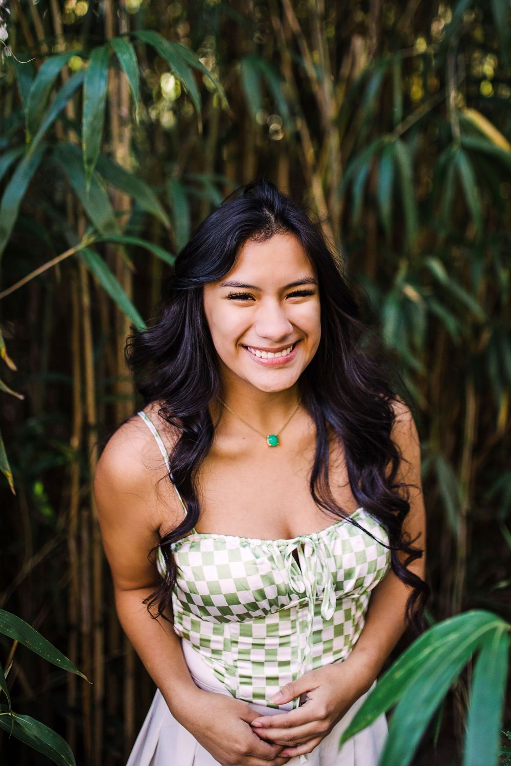 Smiling young woman in green checkered top stands joyfully in front of tall bamboo plants.